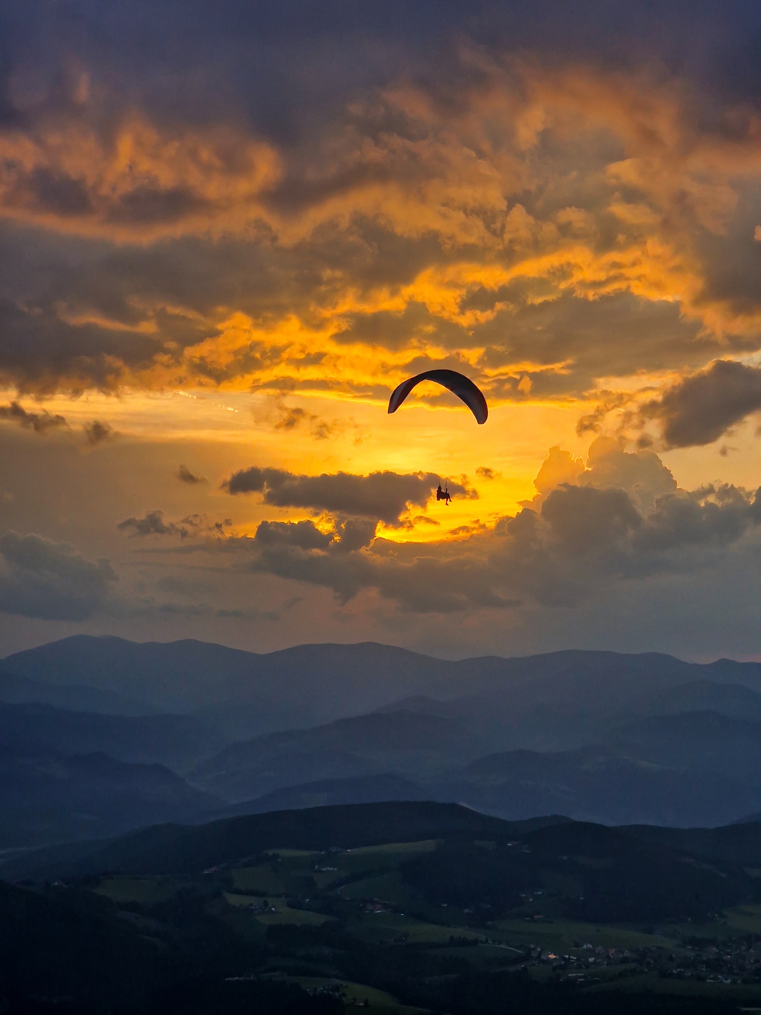 Gleitschirmflieger vor einem dramatischen Sonnenuntergang mit Bergen und Tälern im Hintergrund.
