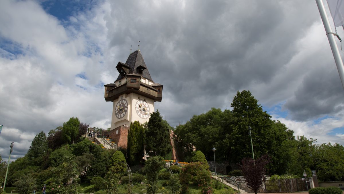 Grazer Uhrturm mit Blumenbeeten im Vordergrund, am Himmel ziehen dunkle Wolken auf.