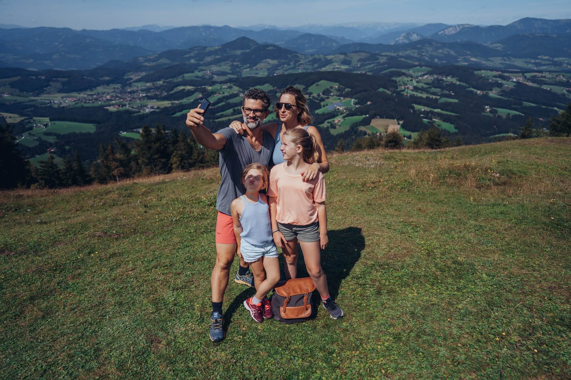Familie mit zwei Mädchen macht am Gipfel des Schöckls ein Selfie, im Hintergrund Grazer Hügelland.