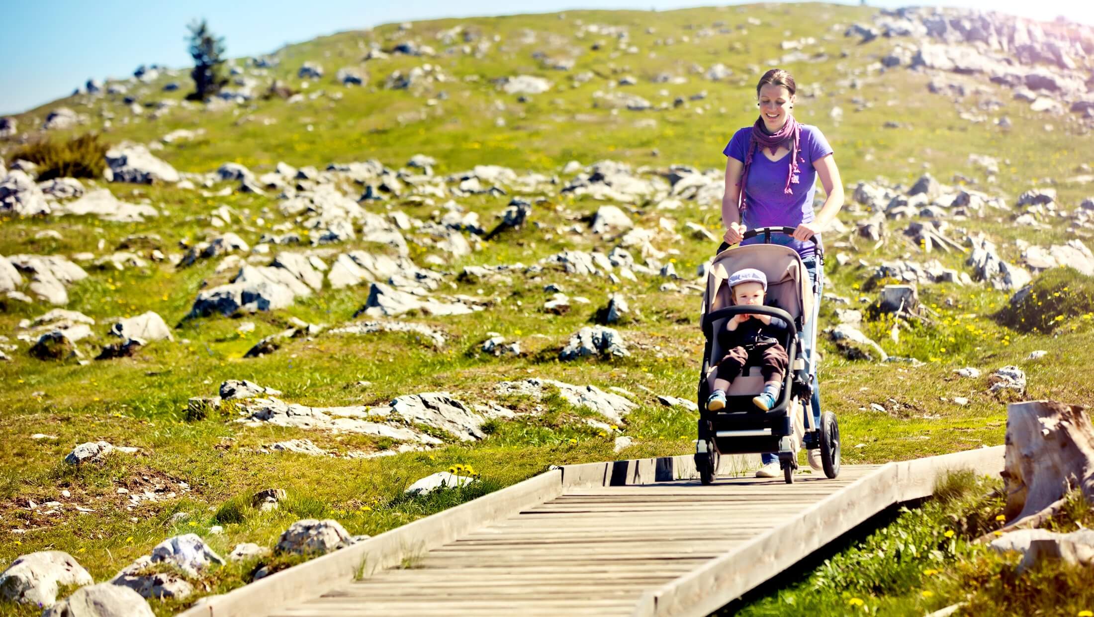 Mutter schiebt Kinderwagen mit Kleinkind am barrierefreien Holzweg am Schöcklplateau.