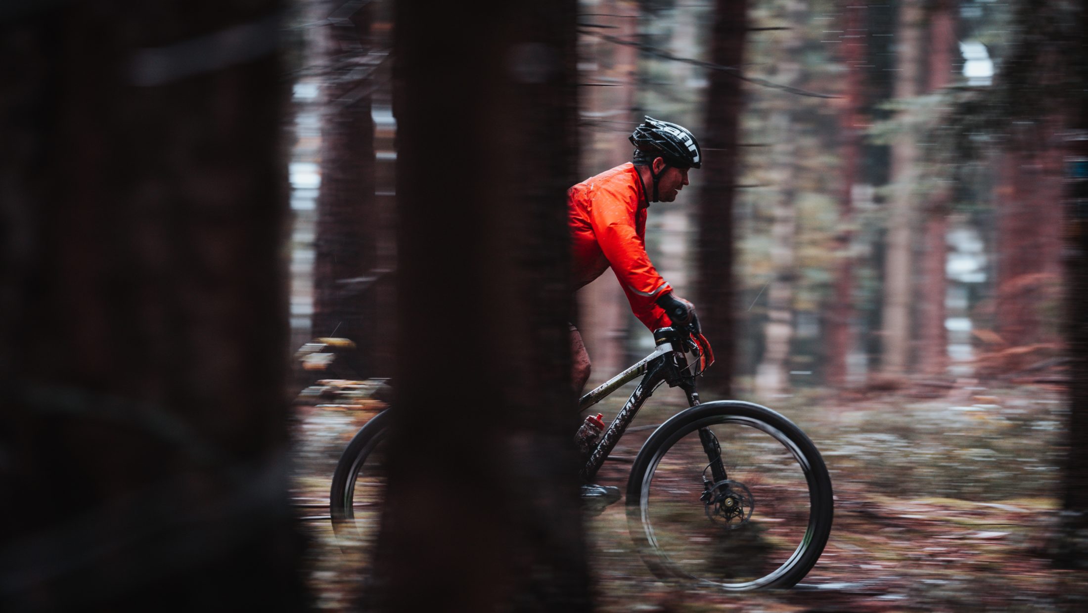 A person in a bright orange jacket rides a bike through a forest. The image is blurred as if taken from a moving vehicle.