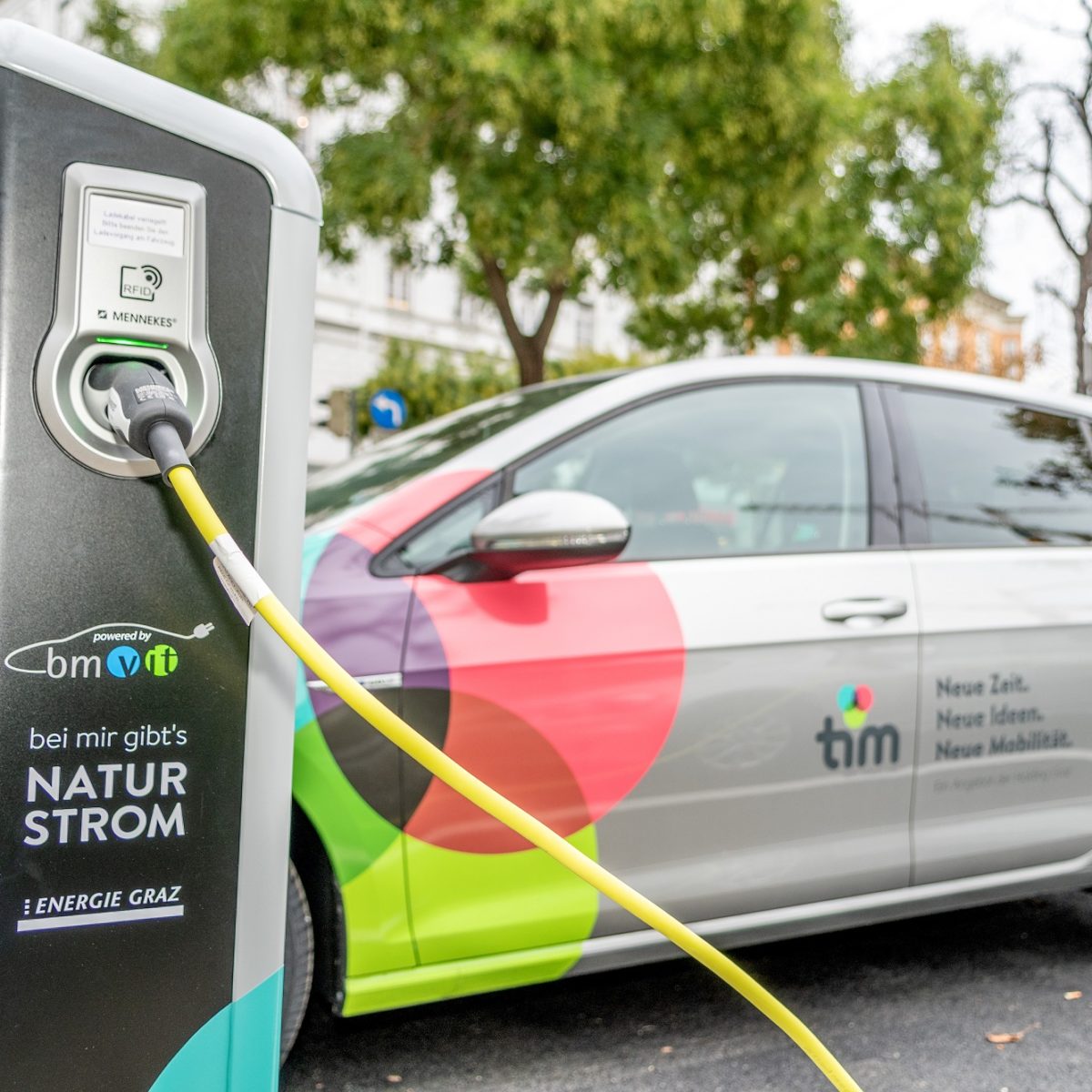 Ladestation mit angeschlossenen Ladekabel, das ein Elektroauto mit Logo von 'tim' auflädt.