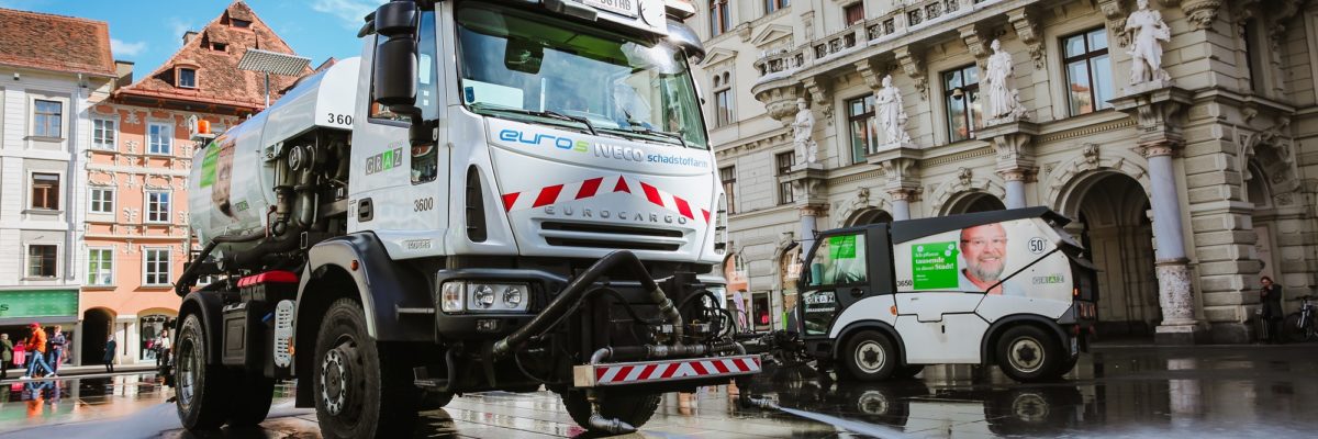 Two street cleaning trucks spray water on the cobblestones in front of the Graz City Hall.