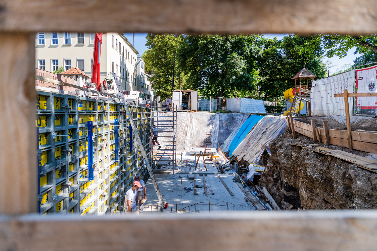 Baustelle mit Schalungswänden, Gerüsten und Bauarbeitern, umgeben von Bäumen und einem Spielplatz