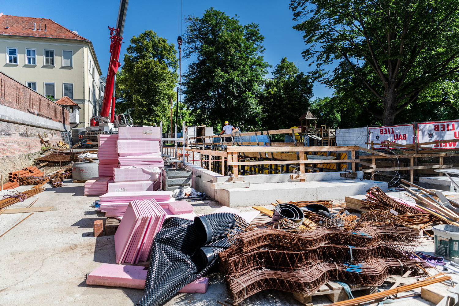 Baustelle mit Baumaterialien, Kran und einem Arbeiter, umgeben von Bäumen und Gebäude.