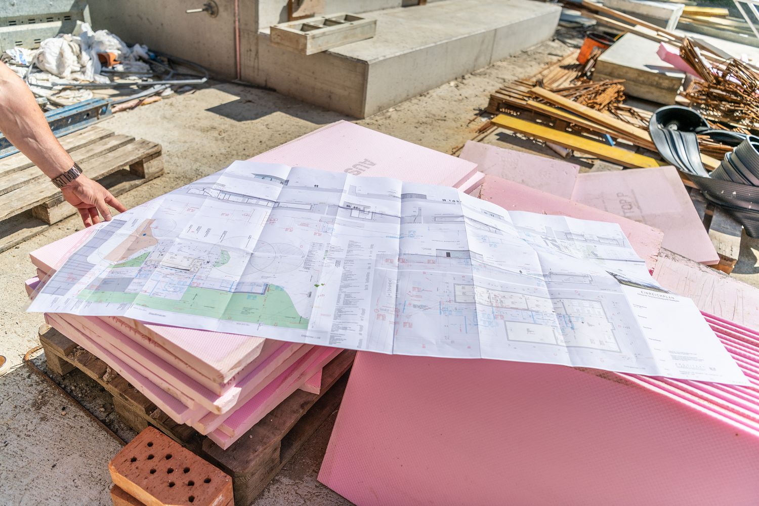 Bauplan auf rosafarbenen Dämmplatten, umgeben von Baumaterialien auf einer Baustelle.
