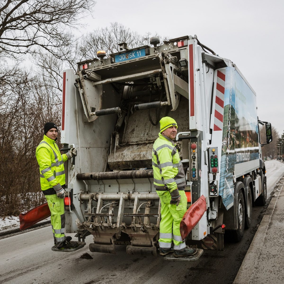 Rubbish truck of Abfallwirtschaft Graz from the rear, two employees in work clothes stand on running boards.
