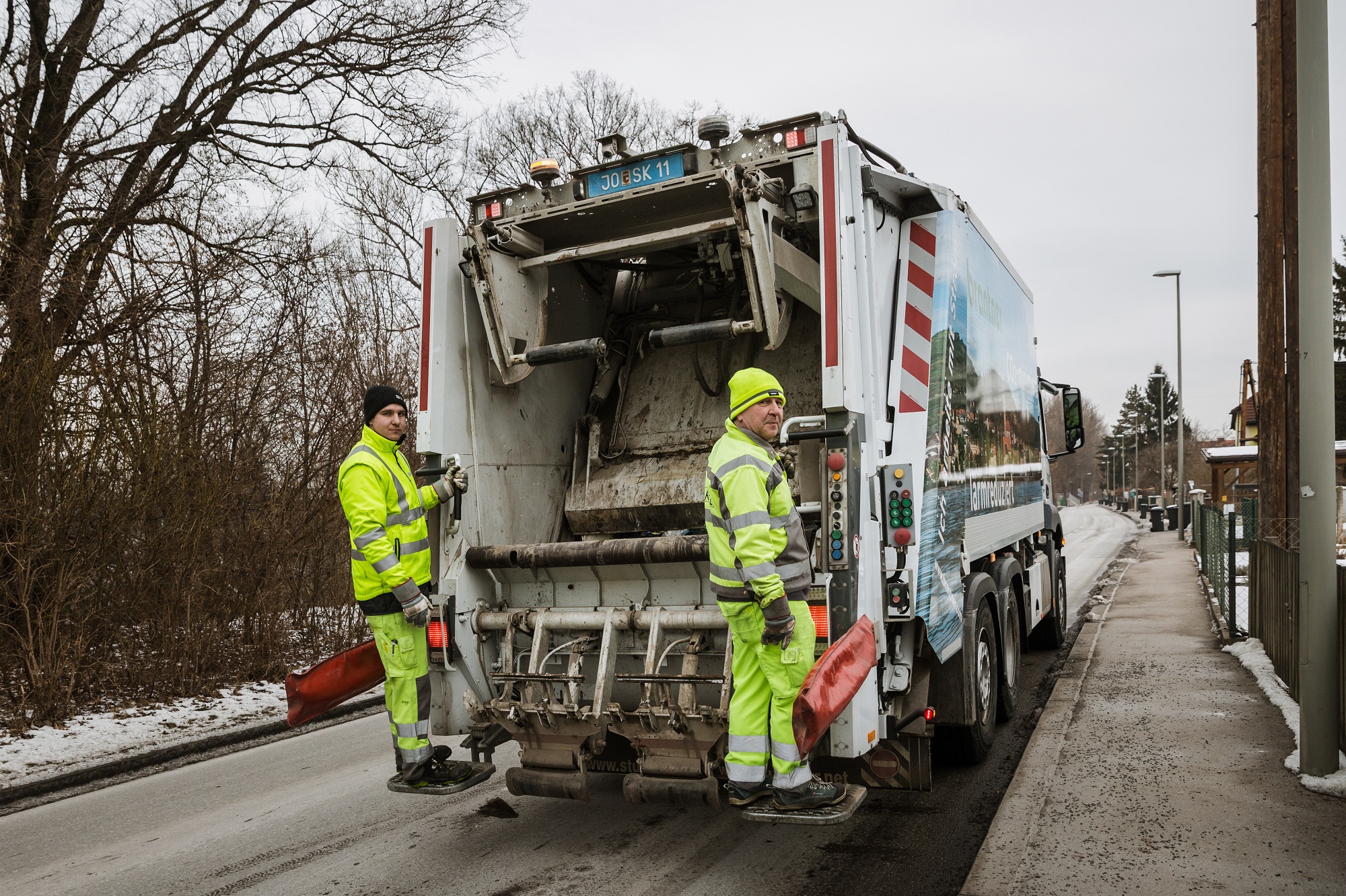 Müllfahrzeug von hinten, auf Trittbrettern stehen zwei Mitarbeiter in Arbeitskleidung