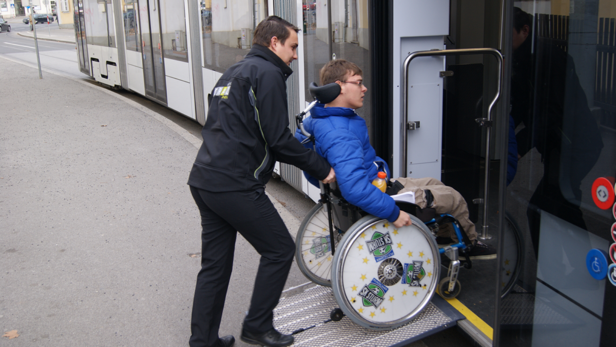 Straßenbahnfahrer schiebt Rollstuhlfahrer über Rampe in einen Bus der Graz Linien