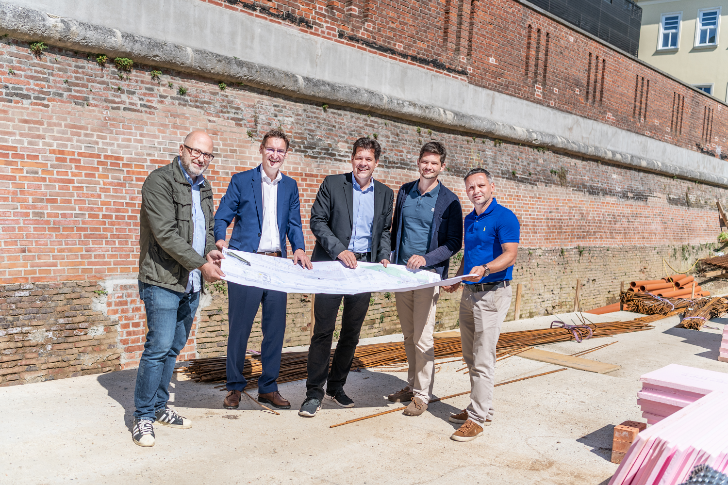 Fünf Männer stehen vor einer Ziegelmauer und betrachten gemeinsam Baupläne auf einer Baustelle