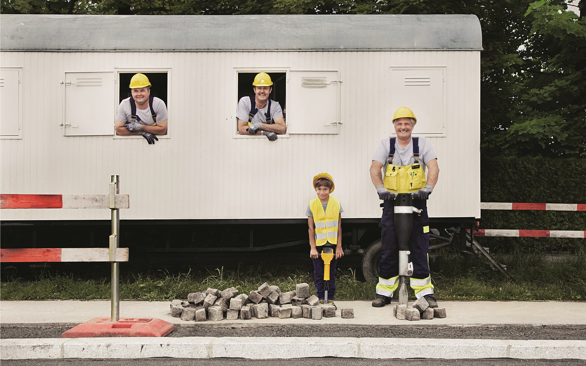 Vier Bauarbeiter, darunter ein Kind, mit gelben Helmen vor einem Baucontainer und Pflastersteinen.