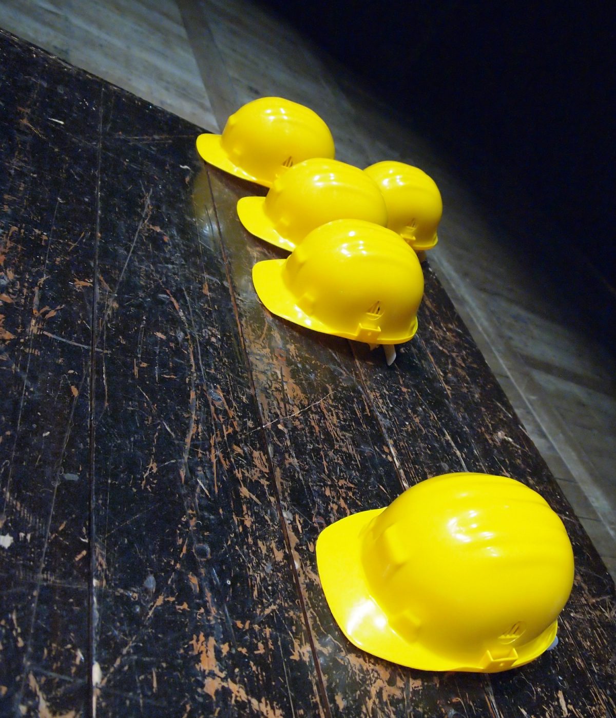 Four yellow hard hats lie in a row on a dark wooden surface.
