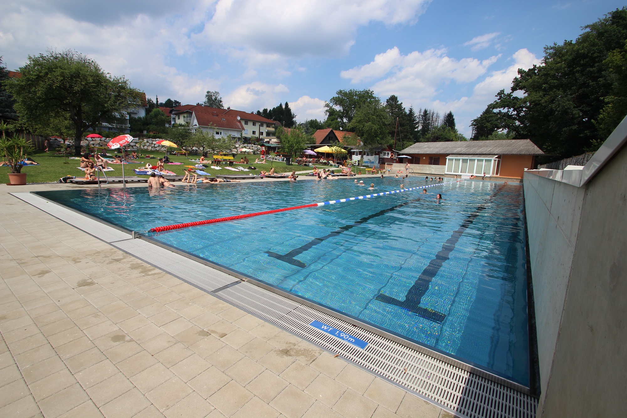 Mehrere Badegäste im Schwimmbecken des Ragnitzbades, vollbesetzte Liegewiese im Hintergrund