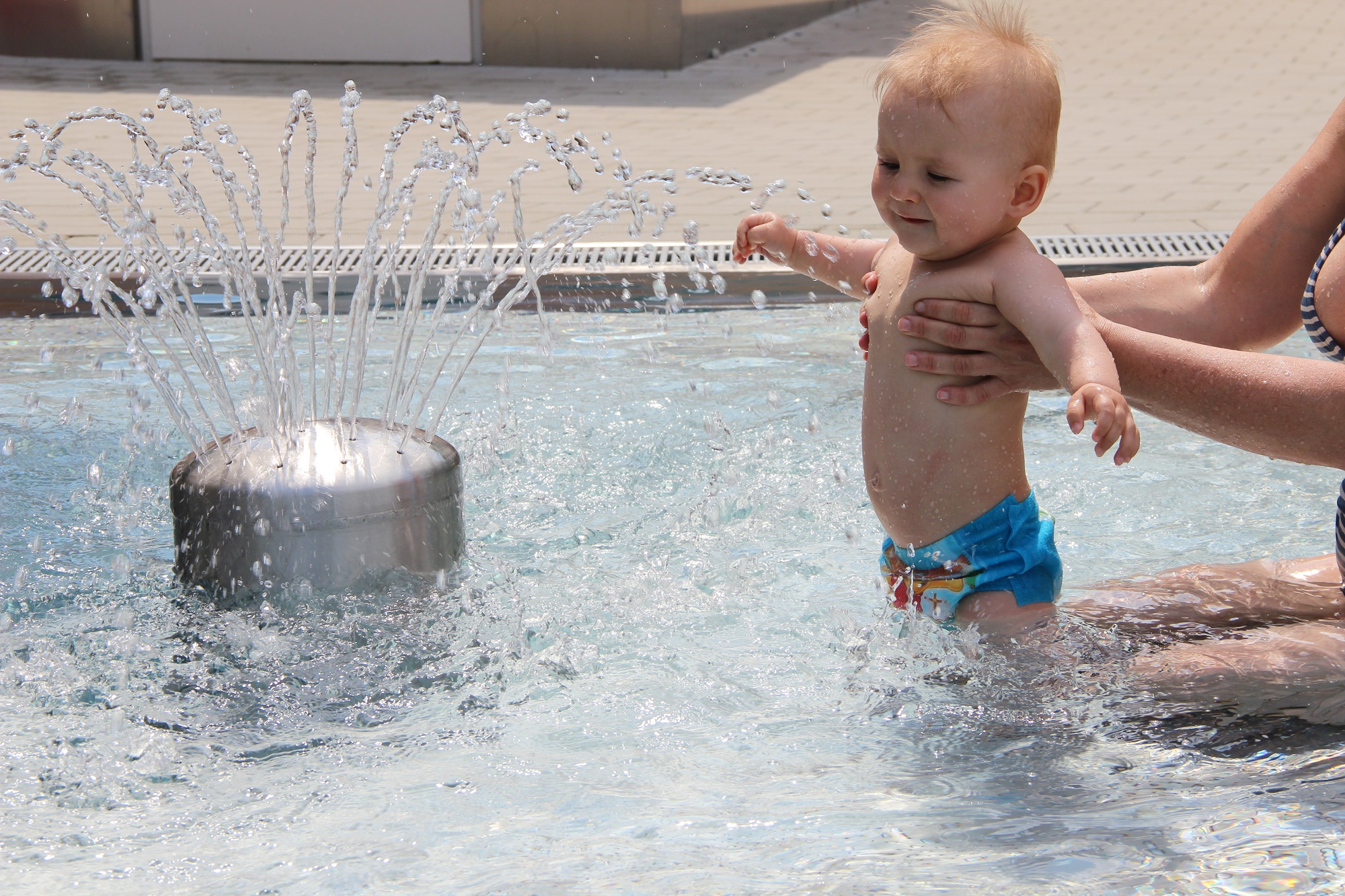 Ragnitzbad: Baby mit Badehose wird von Mutter neben einem sprudelnden Brunnen im Wasser gehalten.
