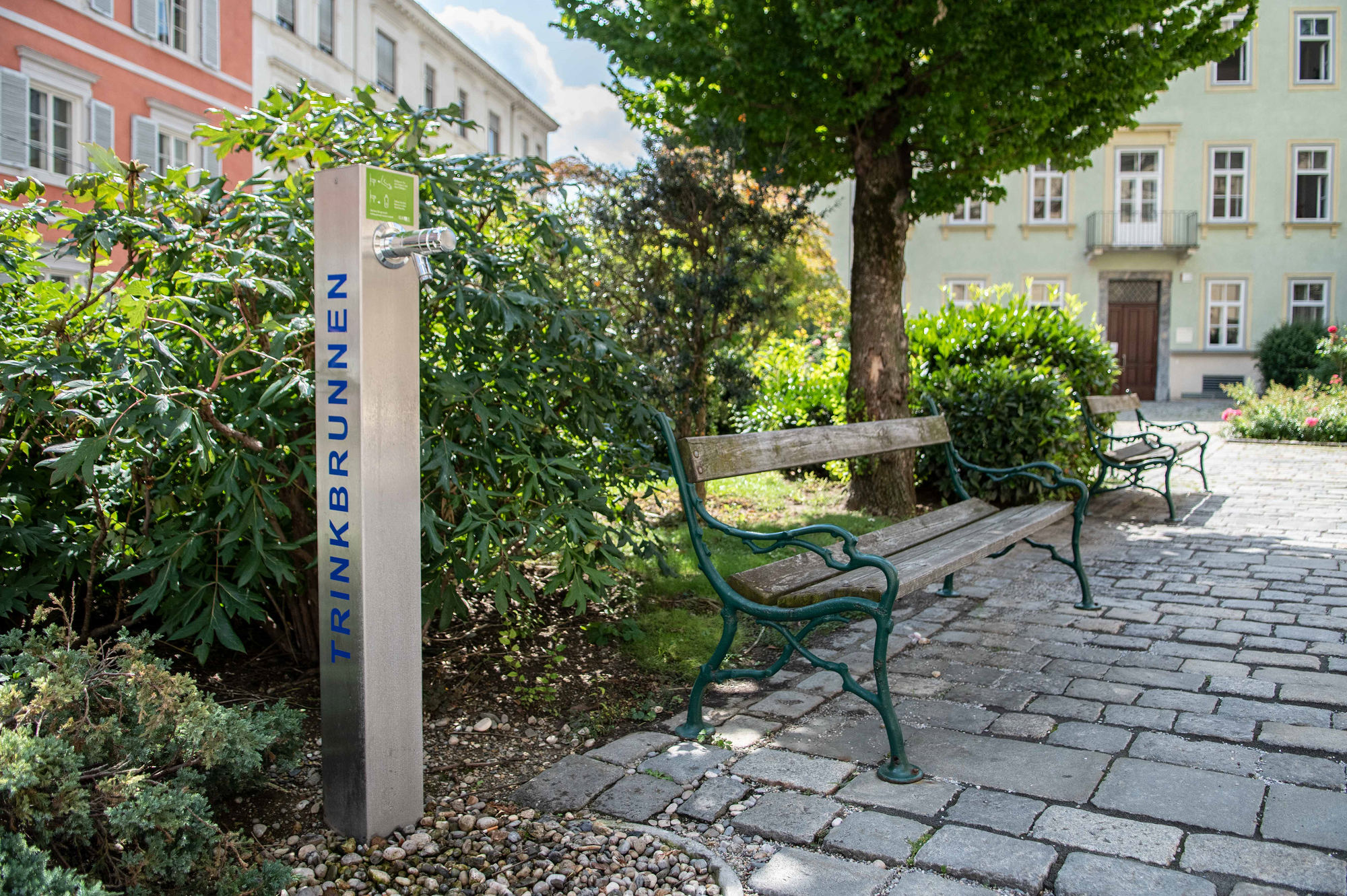 Ein Trinkbrunnen mit der Aufschrift "TRINKBRUNNEN" steht neben zwei Bänken in einer kleinen Grünanlage.