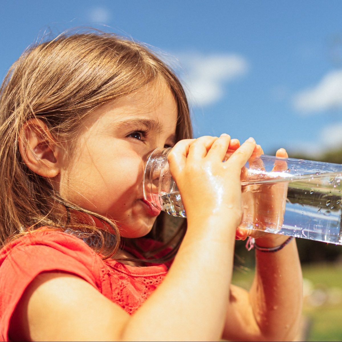 Mädchen in rotem Shirt trinkt bei sonnigem Wetter vor blauem Himmel aus einem Glas Wasser.