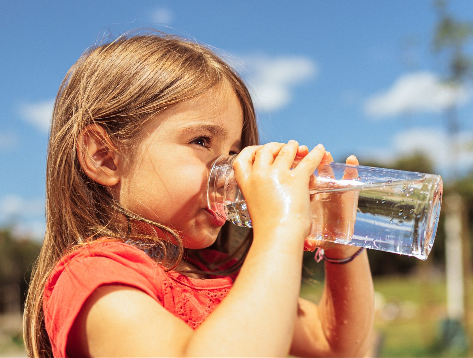 Mädchen in rotem Shirt trinkt bei sonnigem Wetter vor blauem Himmel aus einem Glas Wasser.
