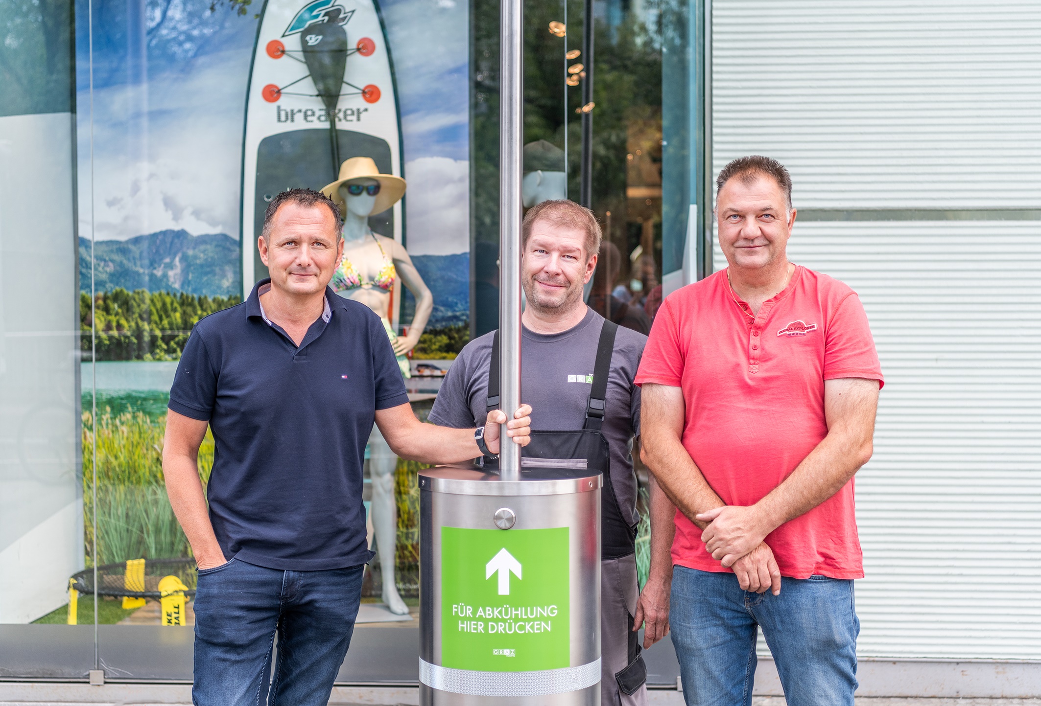 Drei Männer neben Wassersäule mit Aufschrift „Für Abkühlung hier drücken“, Geschäft im Hintergrund.