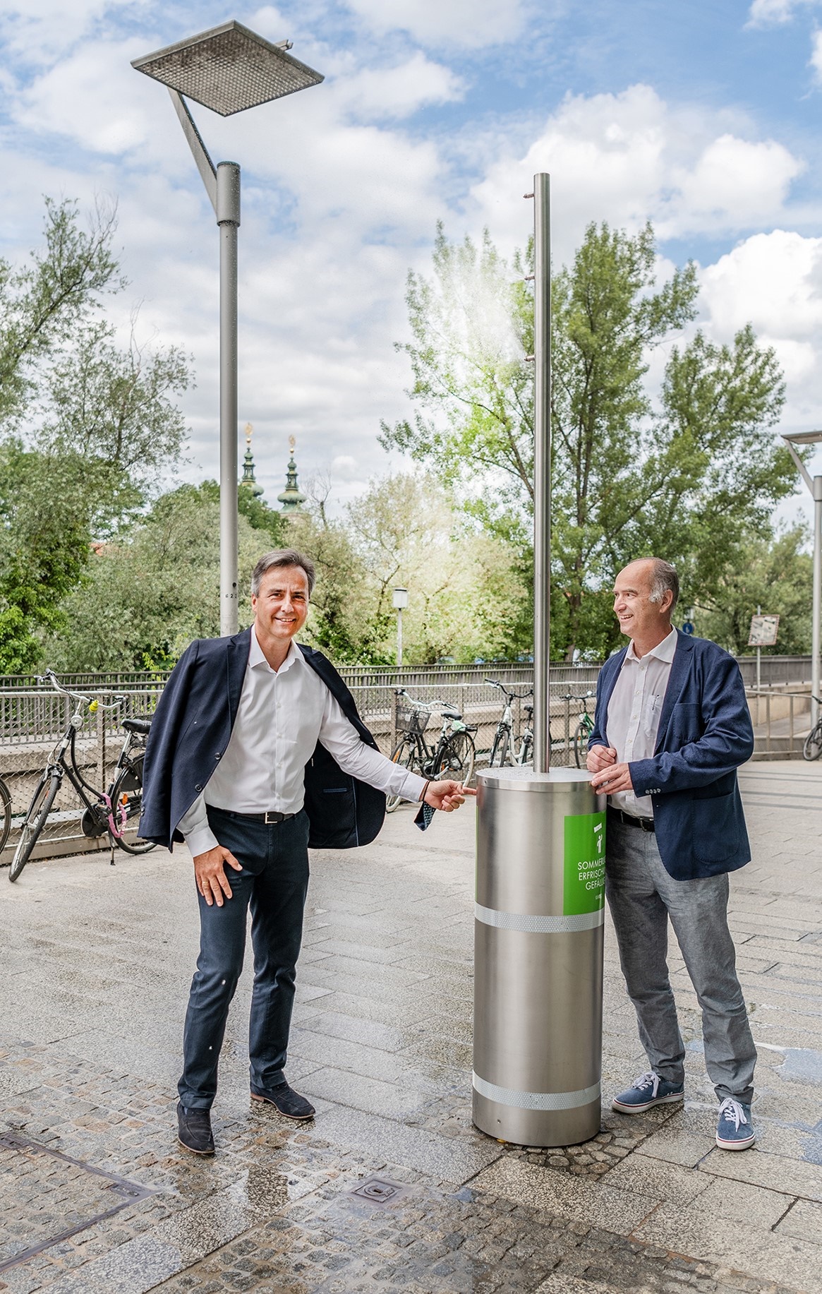Zwei Männer an einer Wassersäule mit Nebel für Erfrischung, Fahrräder und Bäume im Hintergrund.