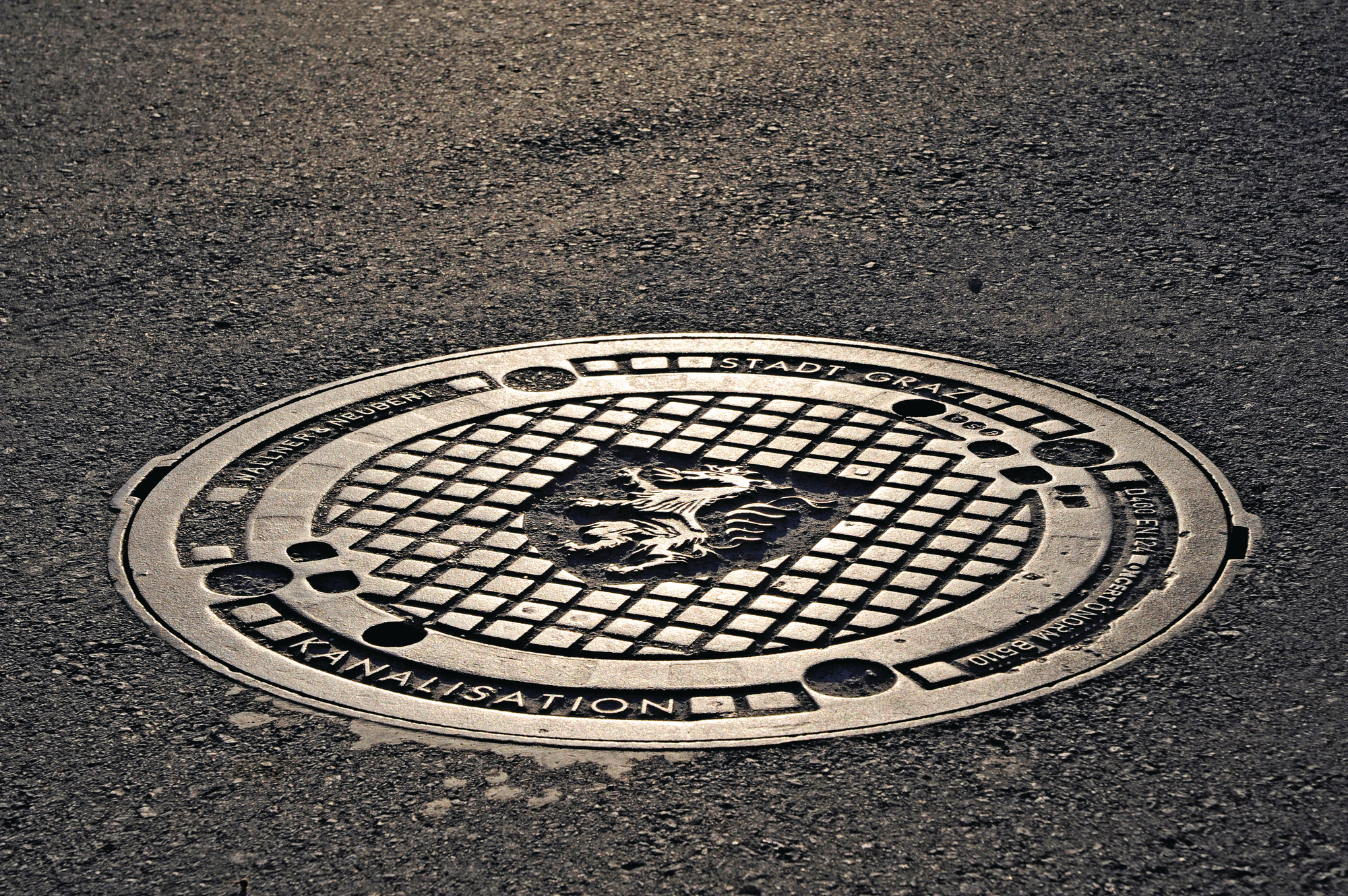 Runder Kanaldeckel der Stadt Graz mit Pantherwappen und Gittermuster auf asphaltierter Straße.