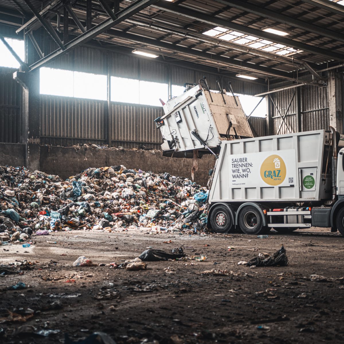 Müllwagen entlädt Abfall in einer Halle, umgeben von großen Müllbergen und industrieller Umgebung.