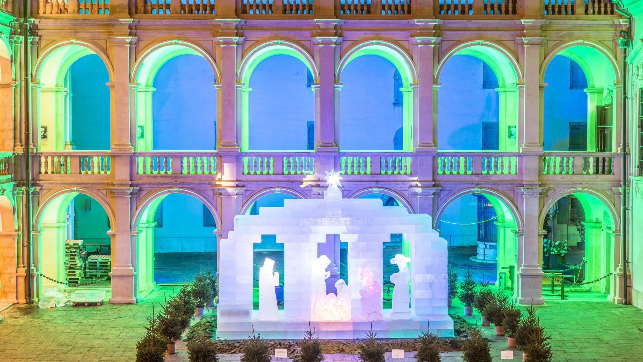 Blick auf die hell erleuchtete Eiskrippe im Landhaushof. Sie ist umringt von kleinen Tannenbäumen.