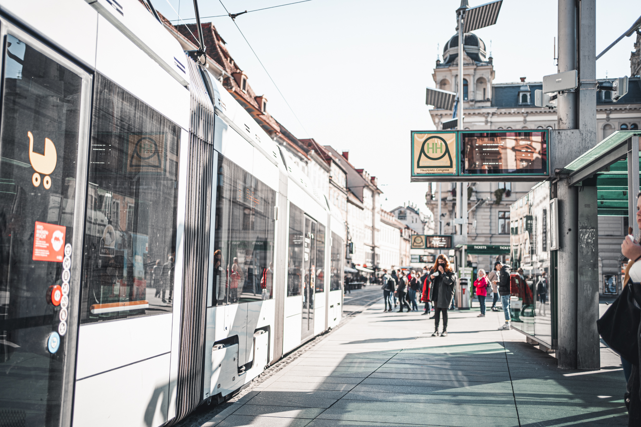 A tram pulls into a stop in Graz, Austria, as people wait on the platform. A sign reads "Hauptbahnhof Congress".