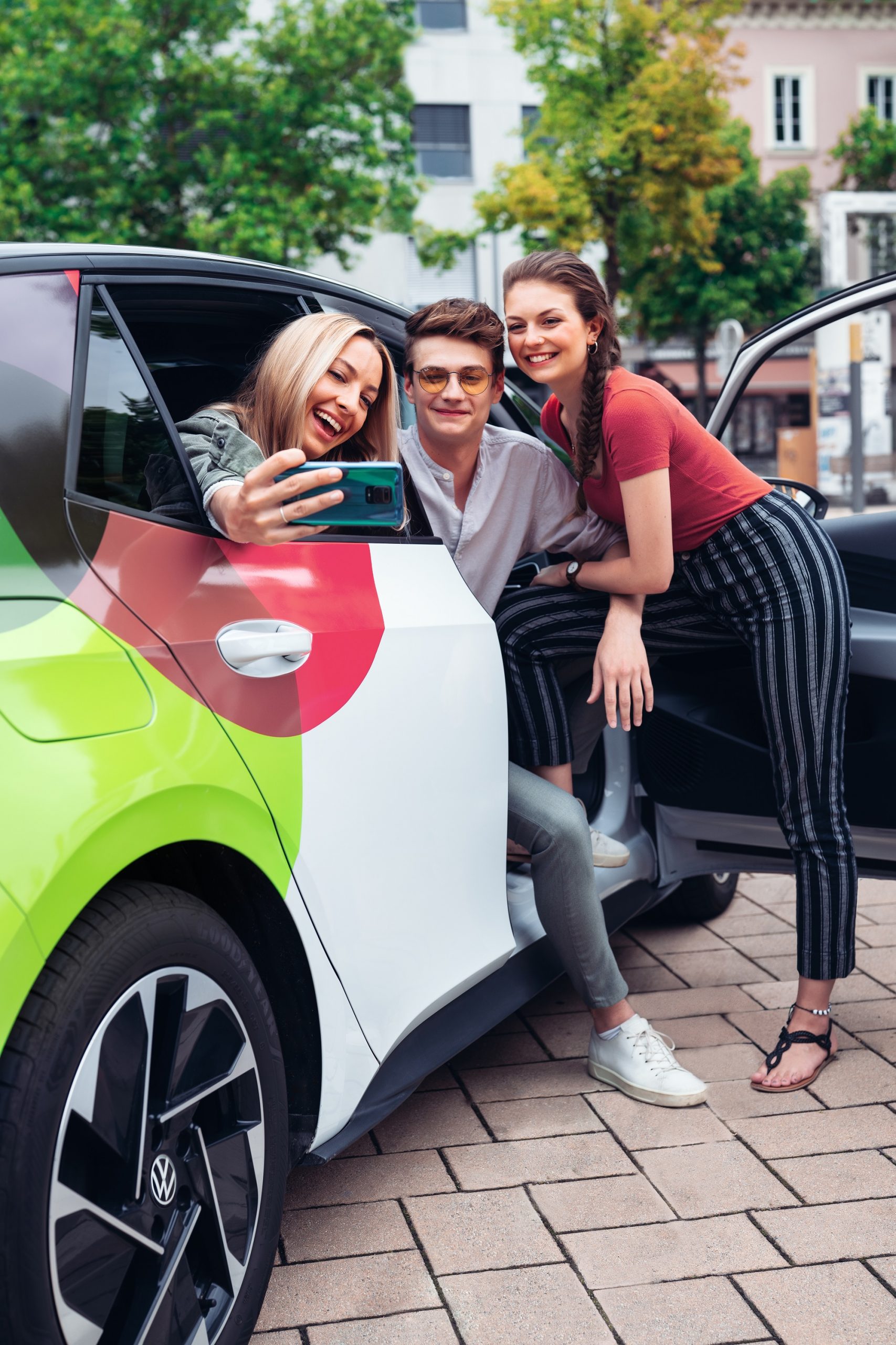 Zwei junge Frauen und ein junger Mann machen mit einem Smartphone ein gemeinsames Selfie an einem tim-carsharing-Auto.