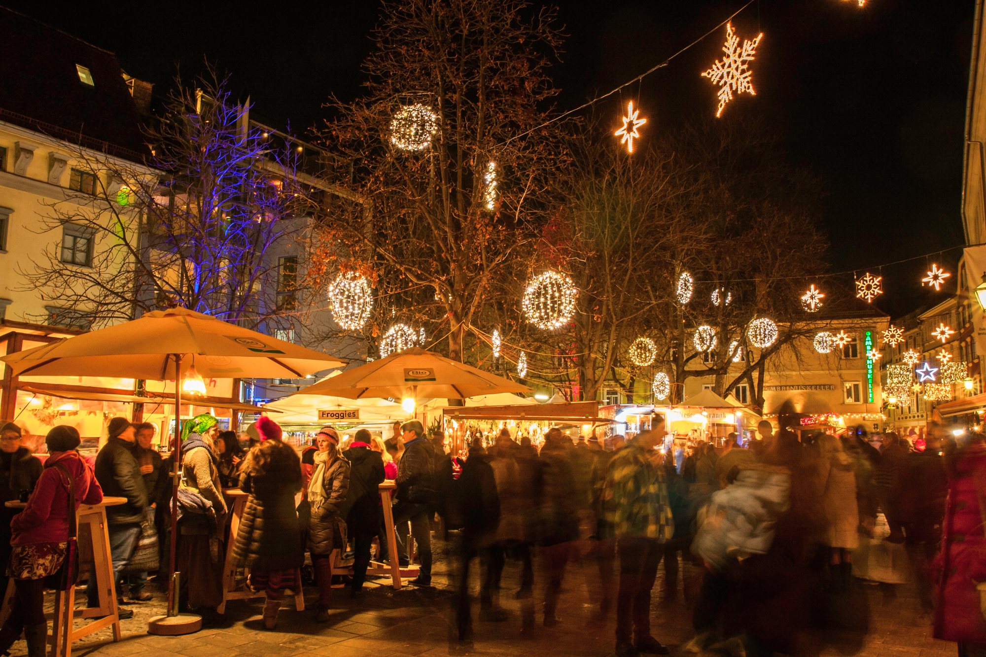 A crowded Christmas market in Graz at night, with people walking around and stalls lit up with fairy lights. "Frogglex" sign visible.
