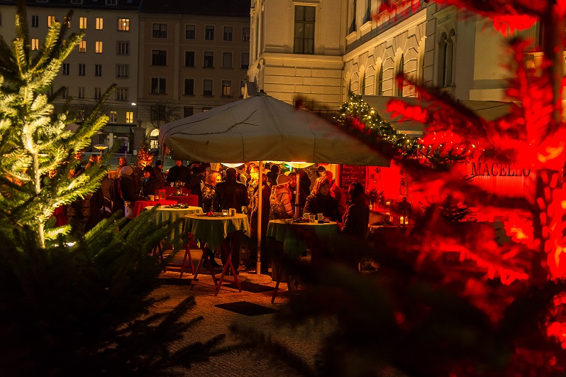 Adventmarkt im Joanneumsviertel mit beleuchteten Ständen, Tannenbäumen und Besuchern bei Nacht.