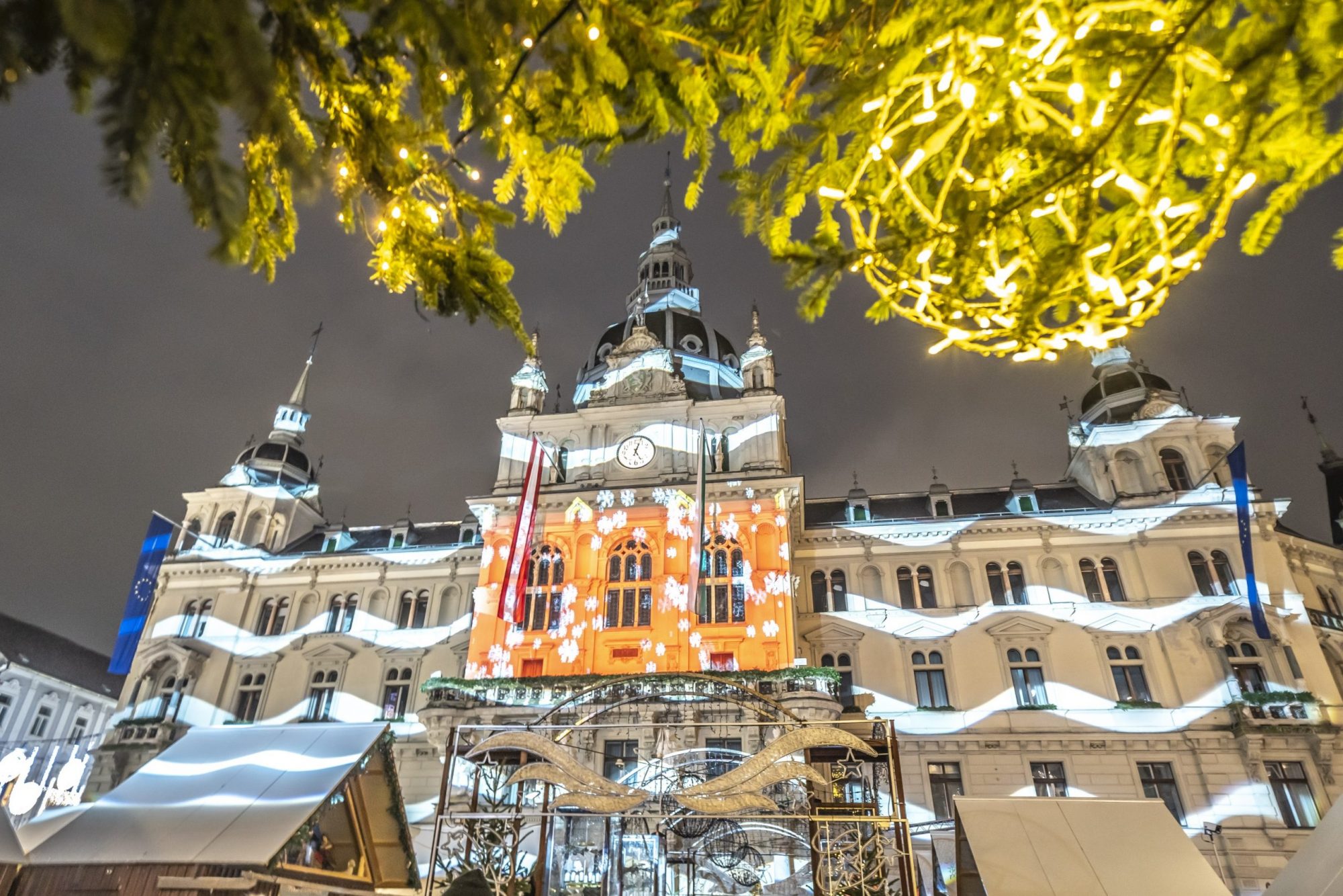 Blick über Adventmarkt auf Grazer Rathaus, auf dessen Fassade weihnachtliche Motive projeziert sind.