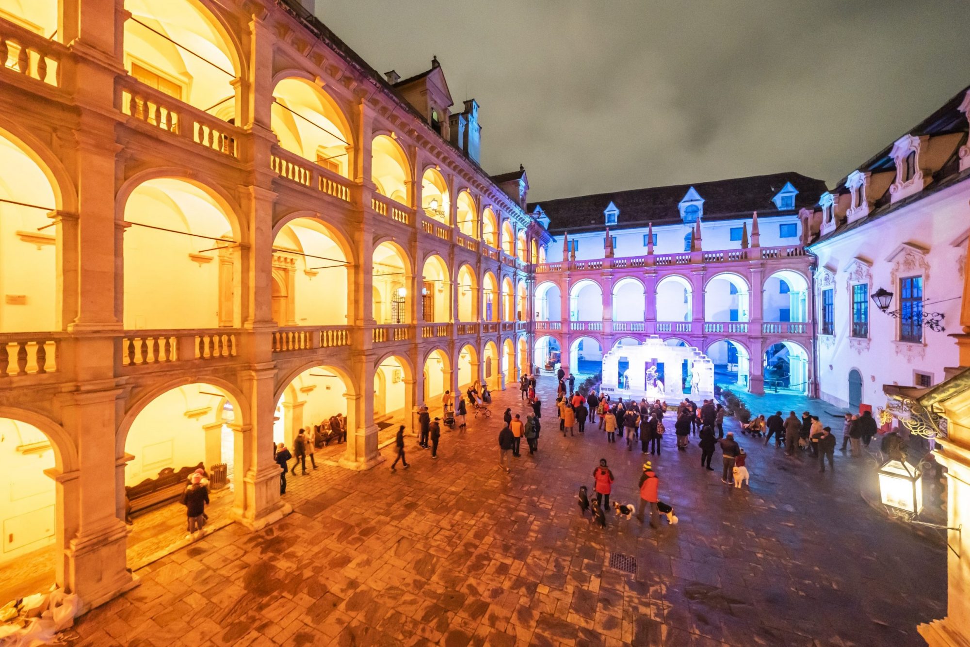 Beleuchteter Landhaushof mit Arkaden, Menschenmenge und einer Eiskrippe im Hintergrund bei Nacht.