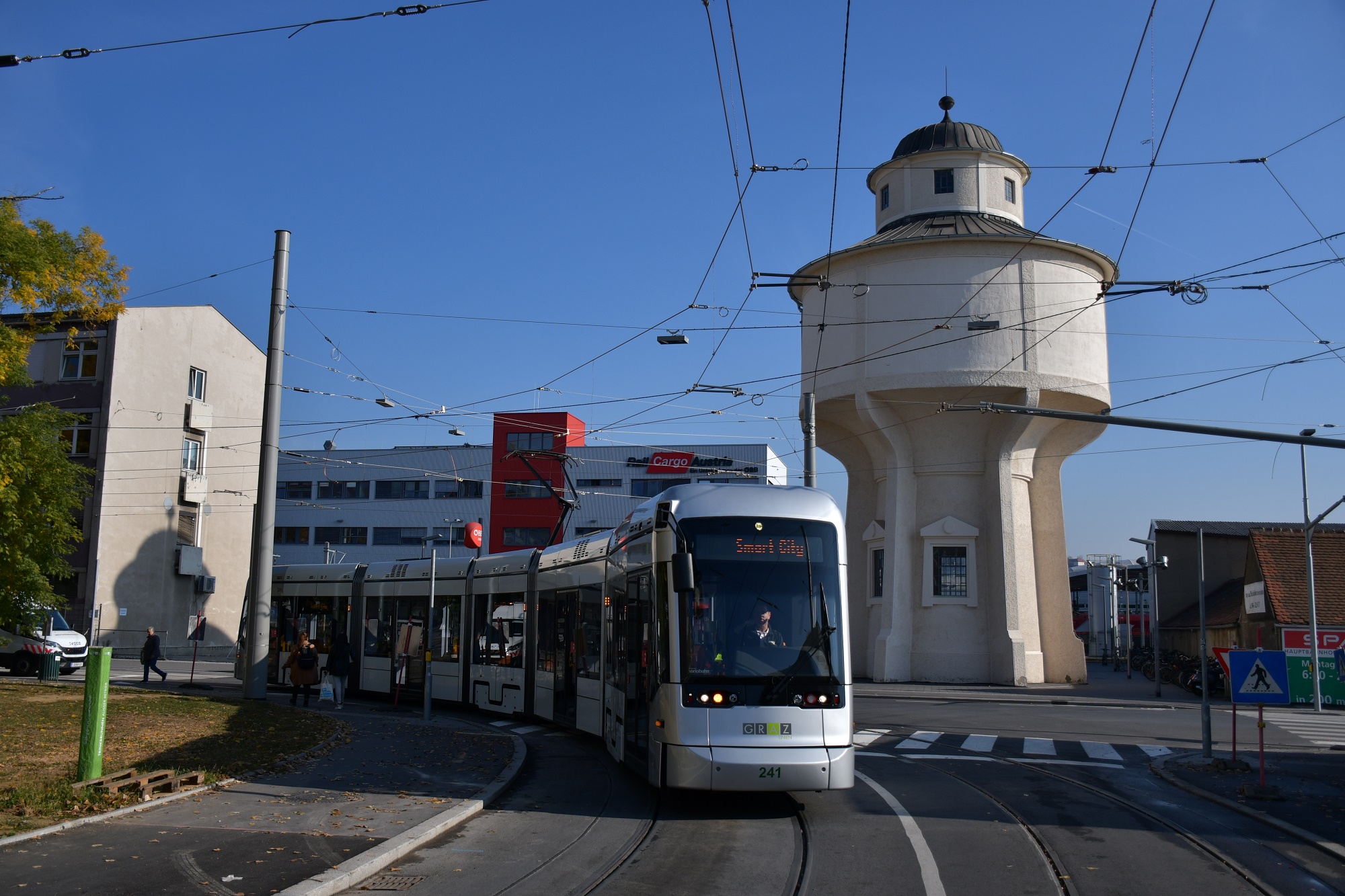Straßenbahn in Graz vor altem Wasserturm, mit klarem Himmel und umliegenden Gebäuden im Hintergrund
