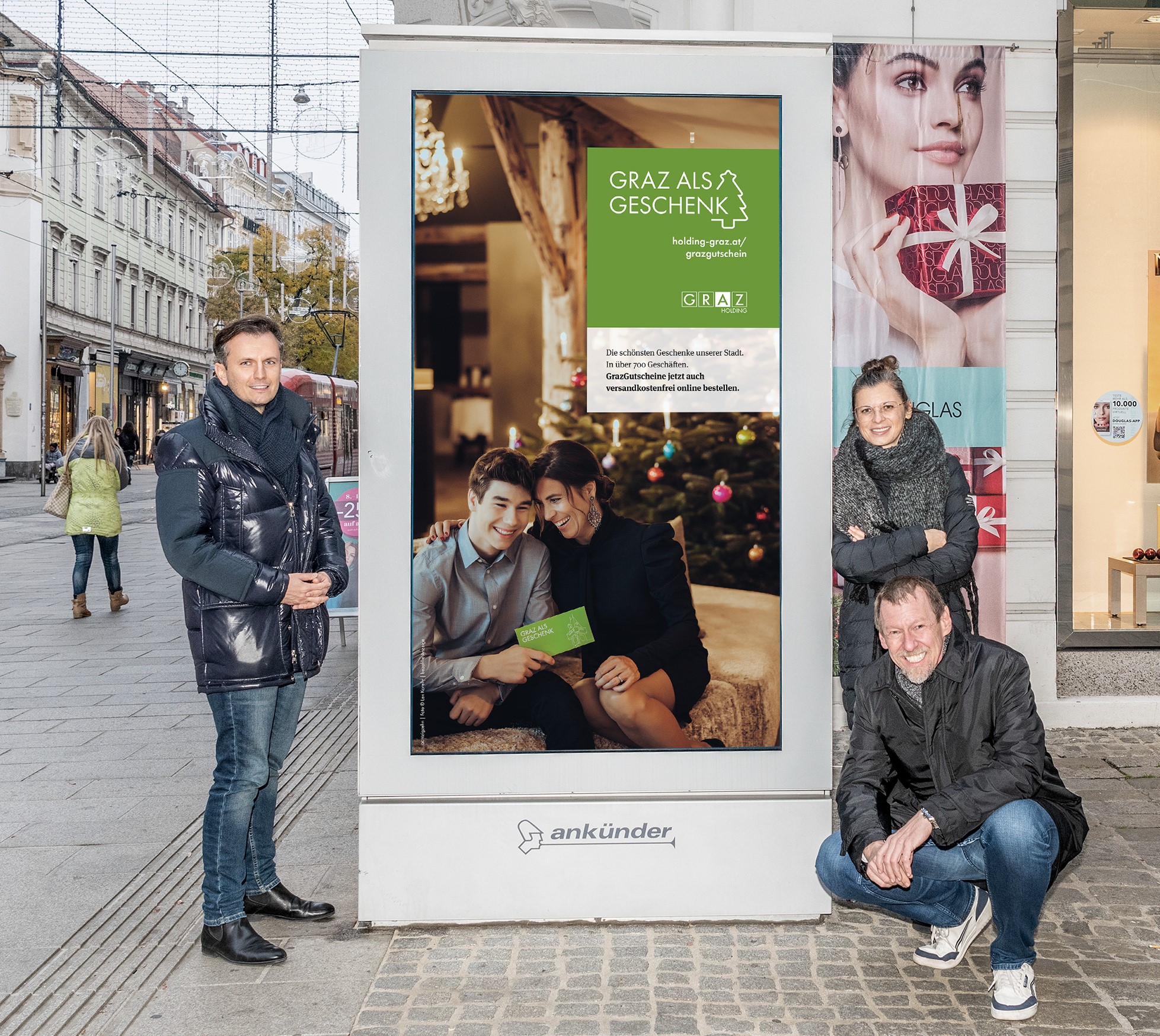 Werbung für GrazGutscheine mit drei Personen vor beleuchteter Infotafel in der Herrengasse.