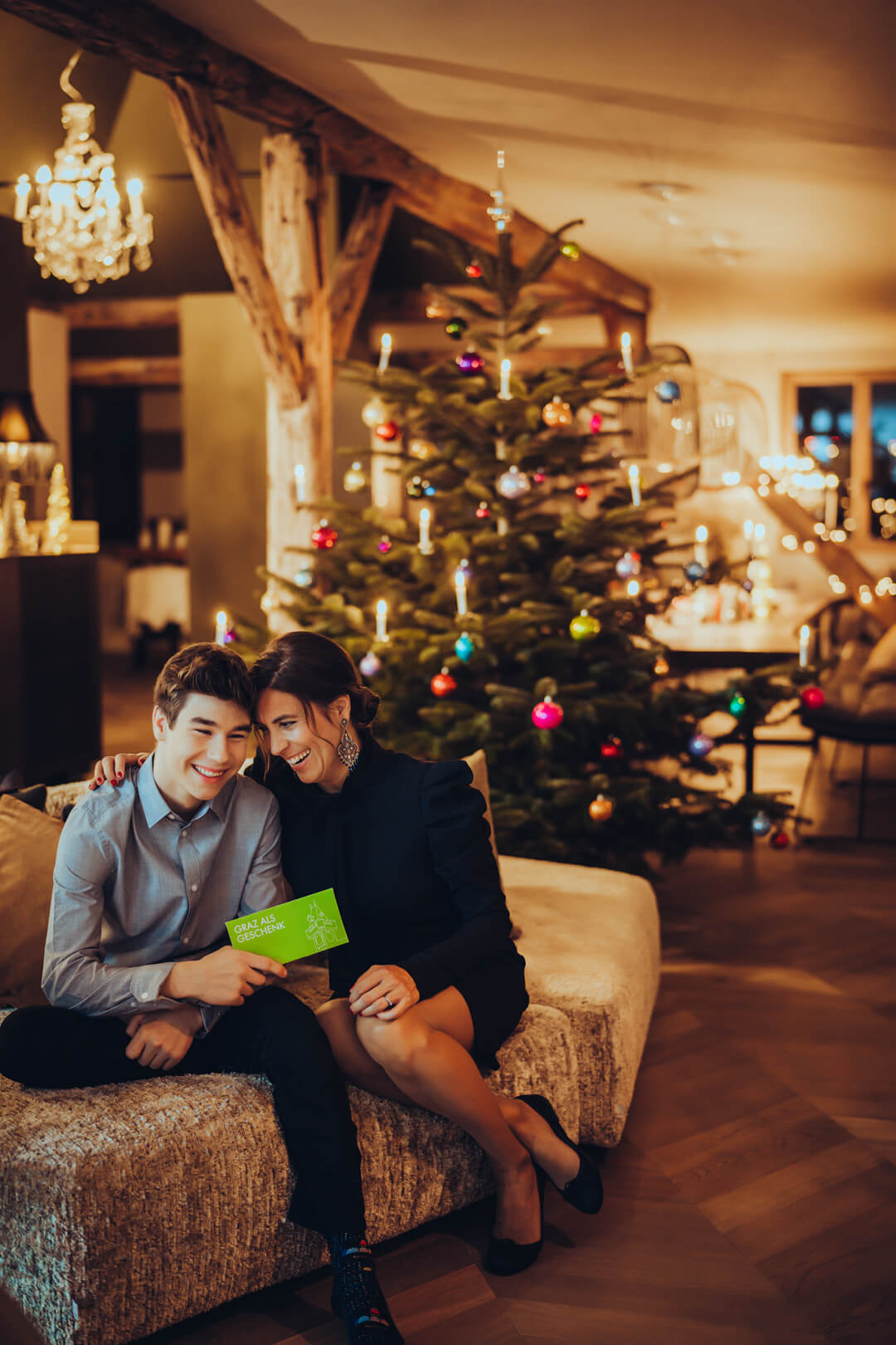Bub und Frau lachen vor festlichem Christbaum und halten grünes Kuvert mit Slogan „Graz als Geschenk“.