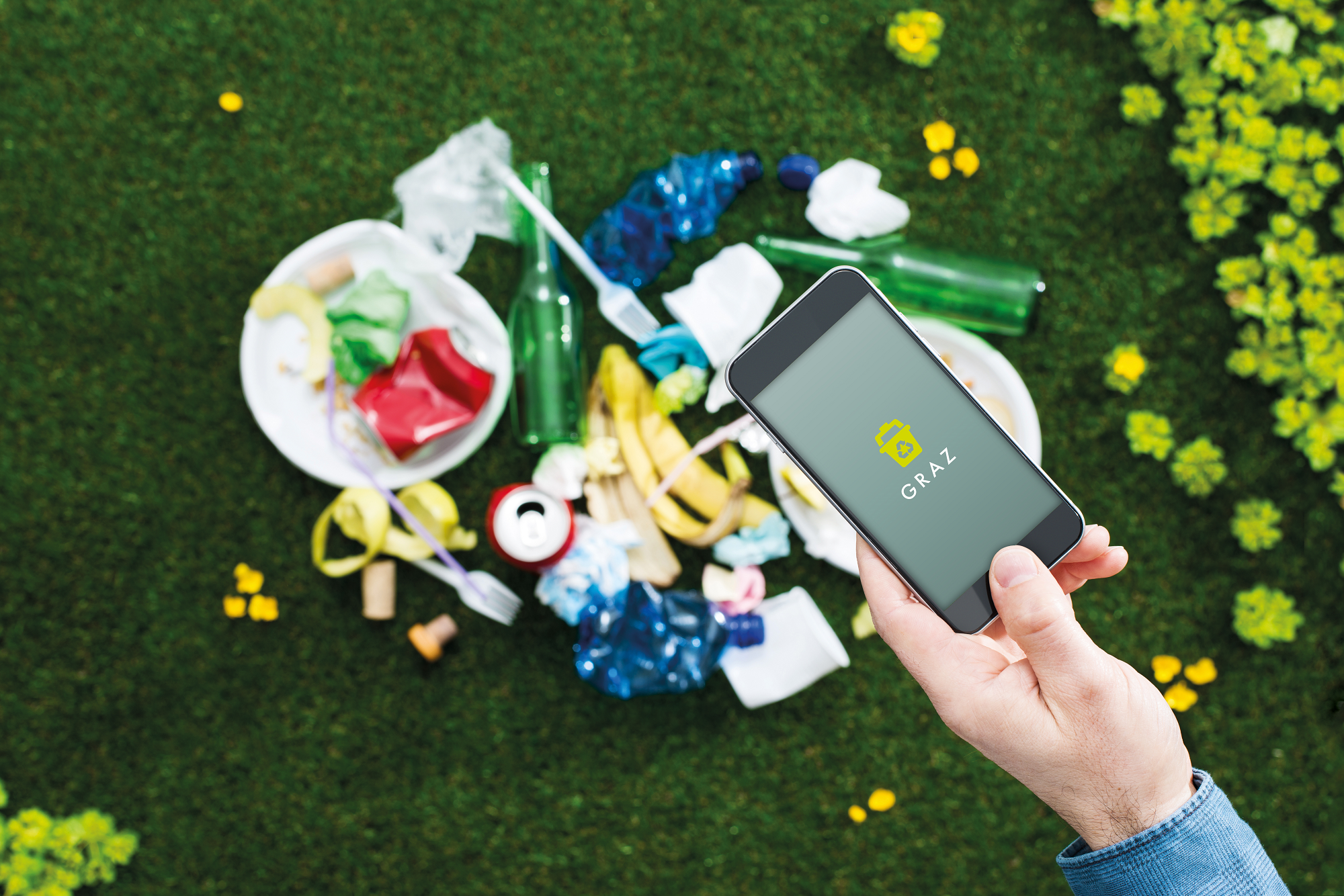 Hand hält Smartphone mit Graz-Logo über verstreutem Müll auf grünem Rasen mit gelben Blüten