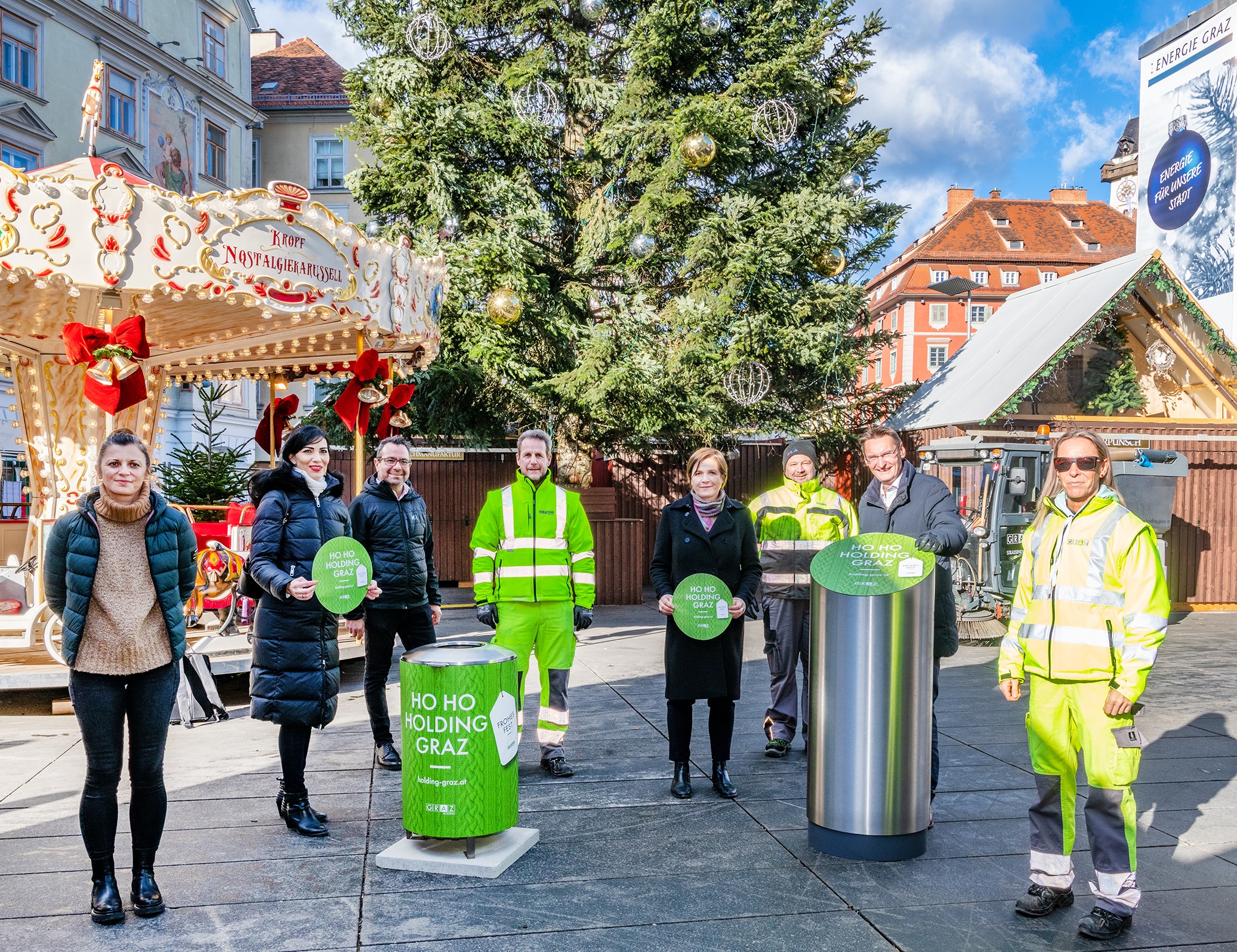 Gruppe vor Weihnachtsbaum am Grazer Hauptplatz präsentiert grüne Mülltonne mit Weihnachtsthema.