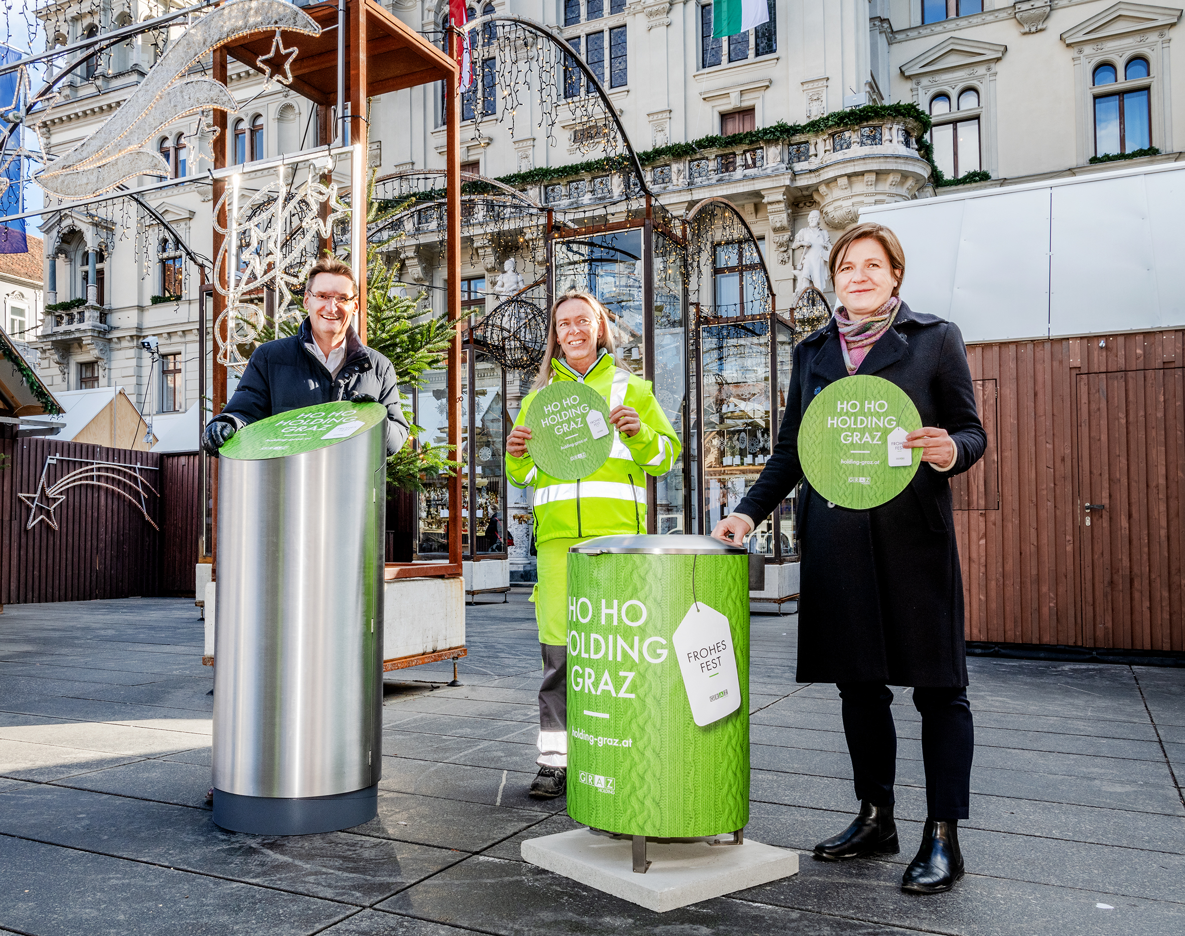 Drei Personen präsentieren weihnachtlich gestaltete Abfallbehälter auf einem Platz vor festlicher Kulisse.