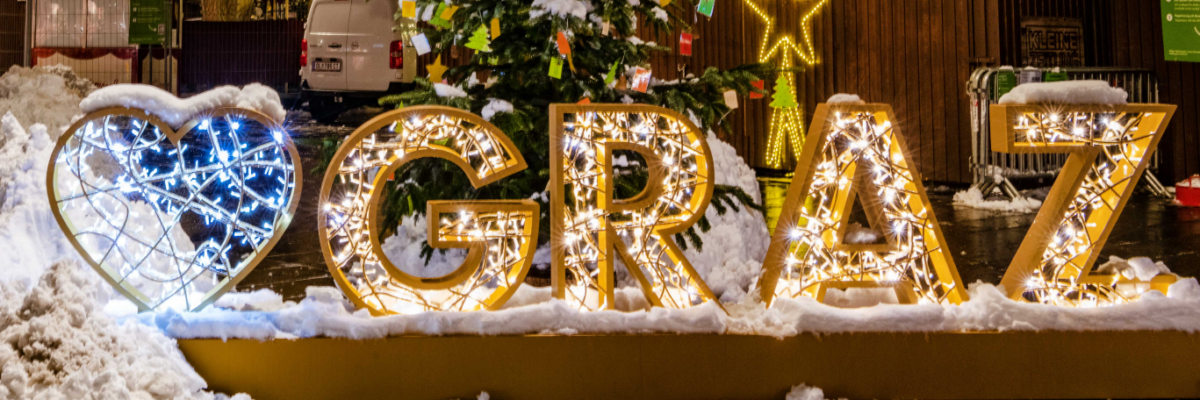 Beleuchteter 'I ❤️ Graz'-Schriftzug mit Schnee bedeckt, Weihnachtsbaum und Stern im Hintergrund.