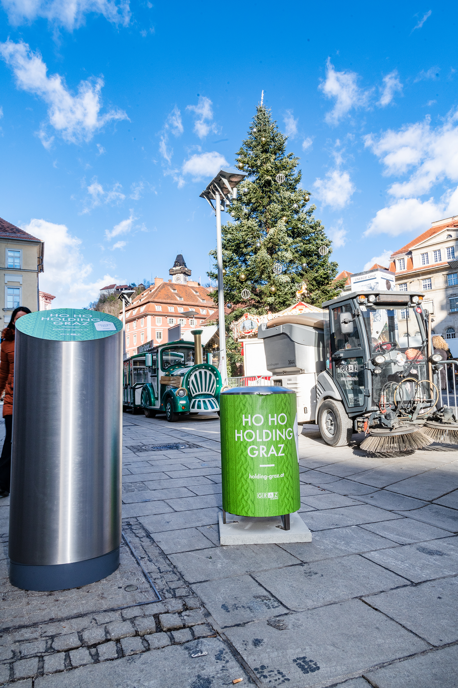 Weihnachtsbaum, Kehrmaschine und Müllbehälter mit Graz-Logo auf Hauptplatz bei blauem Himmel.