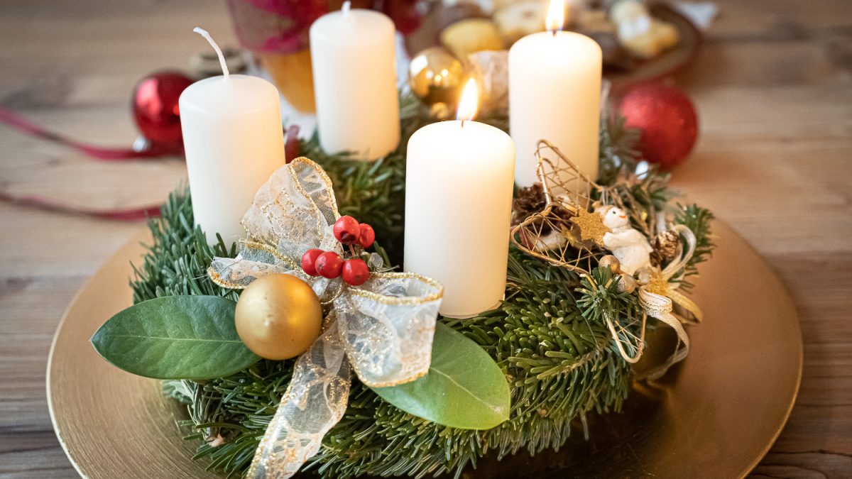 Adventkranz mit weißen Kerzen, goldenen Kugeln, Schleifen und Dekoration auf goldenem Teller.