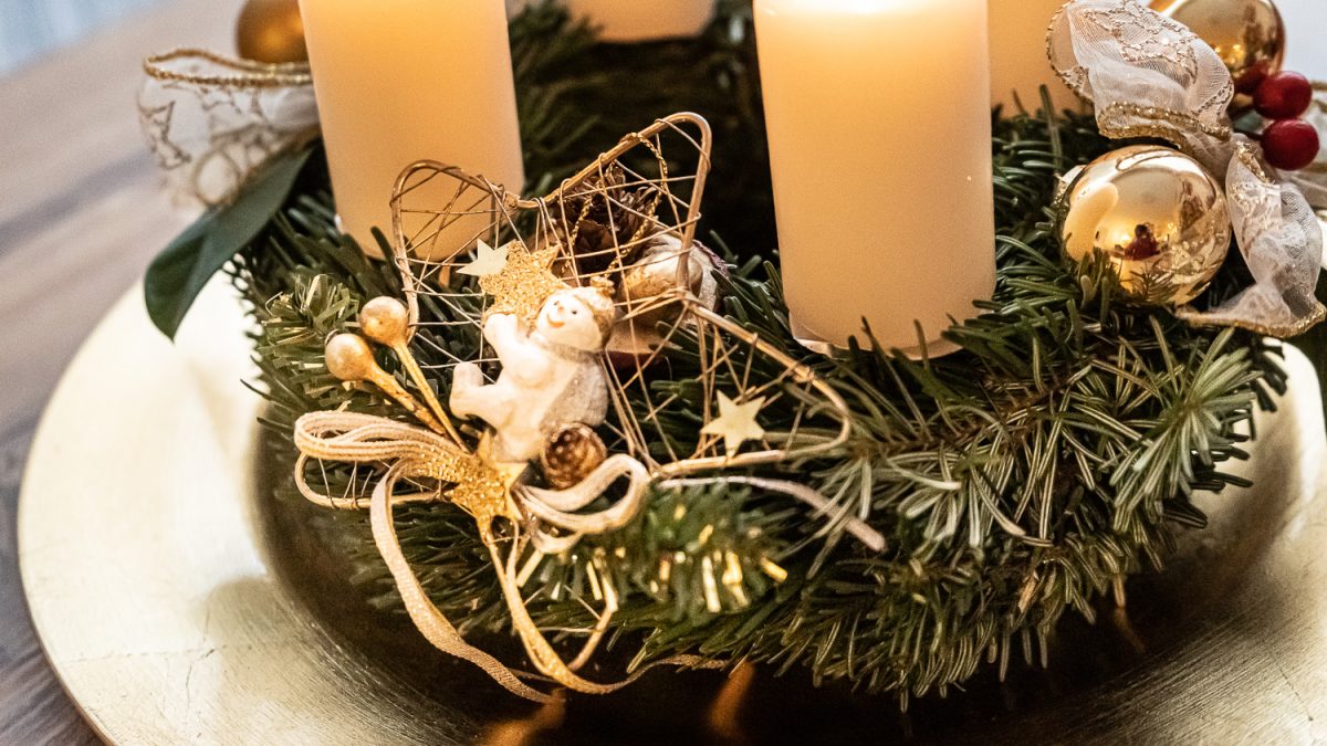 Detail eines Adventkranzes mit weißen Kerzen, goldener Dekoration und Schleifen auf goldenem Teller.