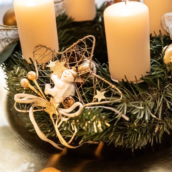 Detail eines Adventkranzes mit weißen Kerzen, goldener Dekoration und Schleifen auf goldenem Teller.