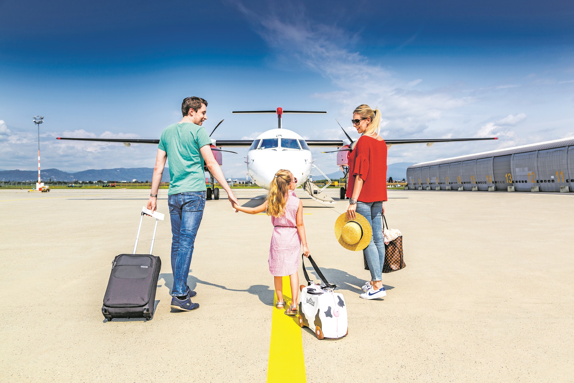 Mutter, Vater und Tochter gehen mit Handgepäck über Rollfeld auf Flugzeug am Flughafen Graz zu.