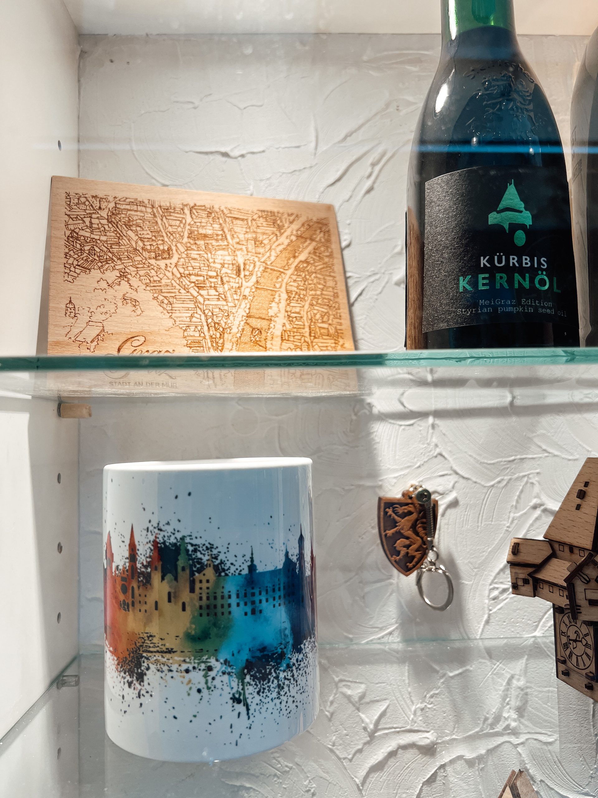 Holzpostkarte mit Abbildung von Graz von oben neben einem steirischen Kürbiskernöl, darunter eine weiße Tasse mit der Skyline von Graz in bunt, Schlüsselanhänger mit steirischem Panther aus Holz und zum Teil sichtbar ein Uhrturm aus Holz in einem Schaufenster