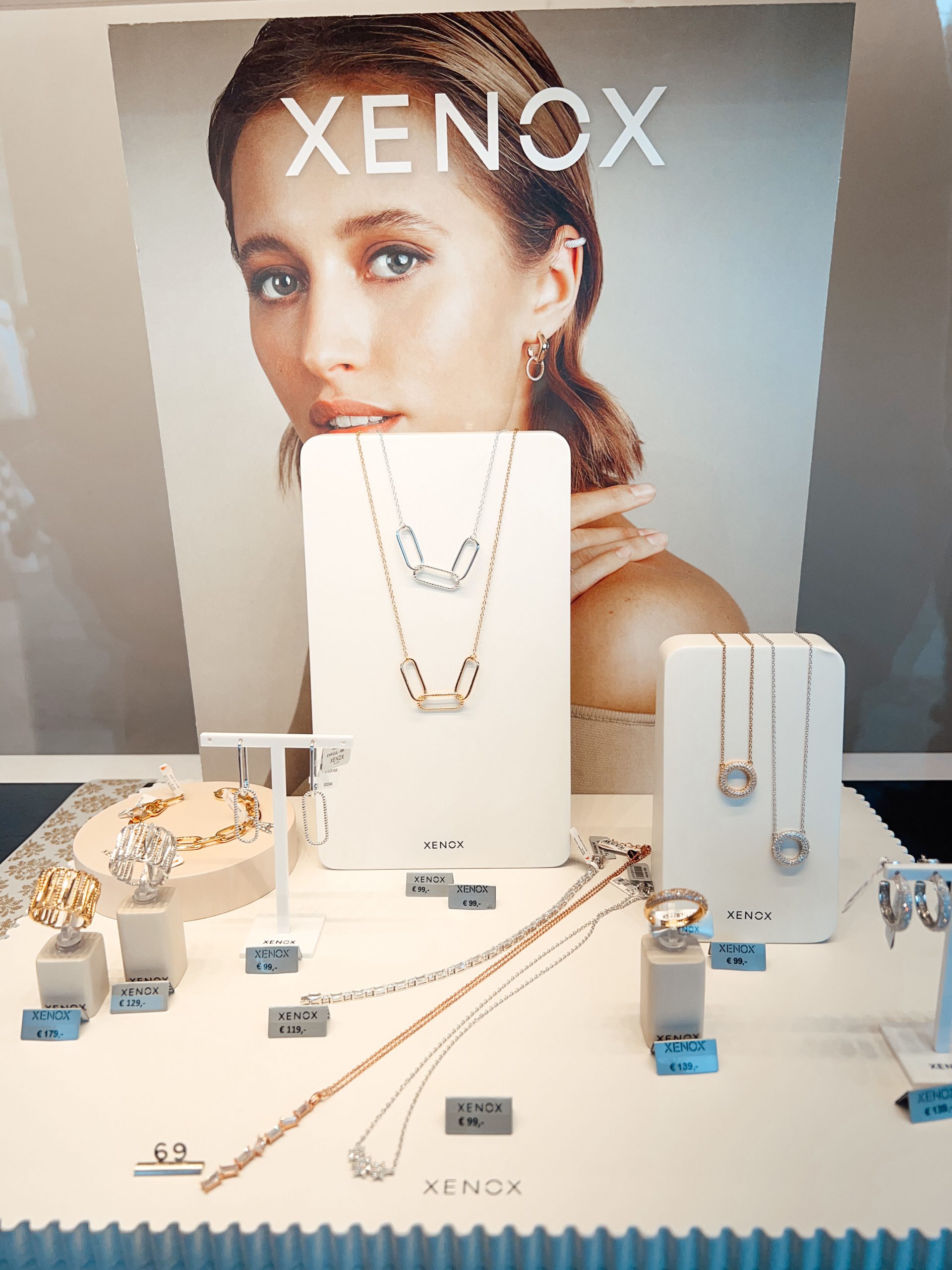Glaskasten mit zwei Paar verschiedenen Ohrringen auf zwei Ständern und mehreren Halsketten und Ringe auf mehreren Ständern, außerdem Ketten und Preisschilder liegend, im Hintergrund ein Werbeplakat der Marke Xenox mit einer Frau