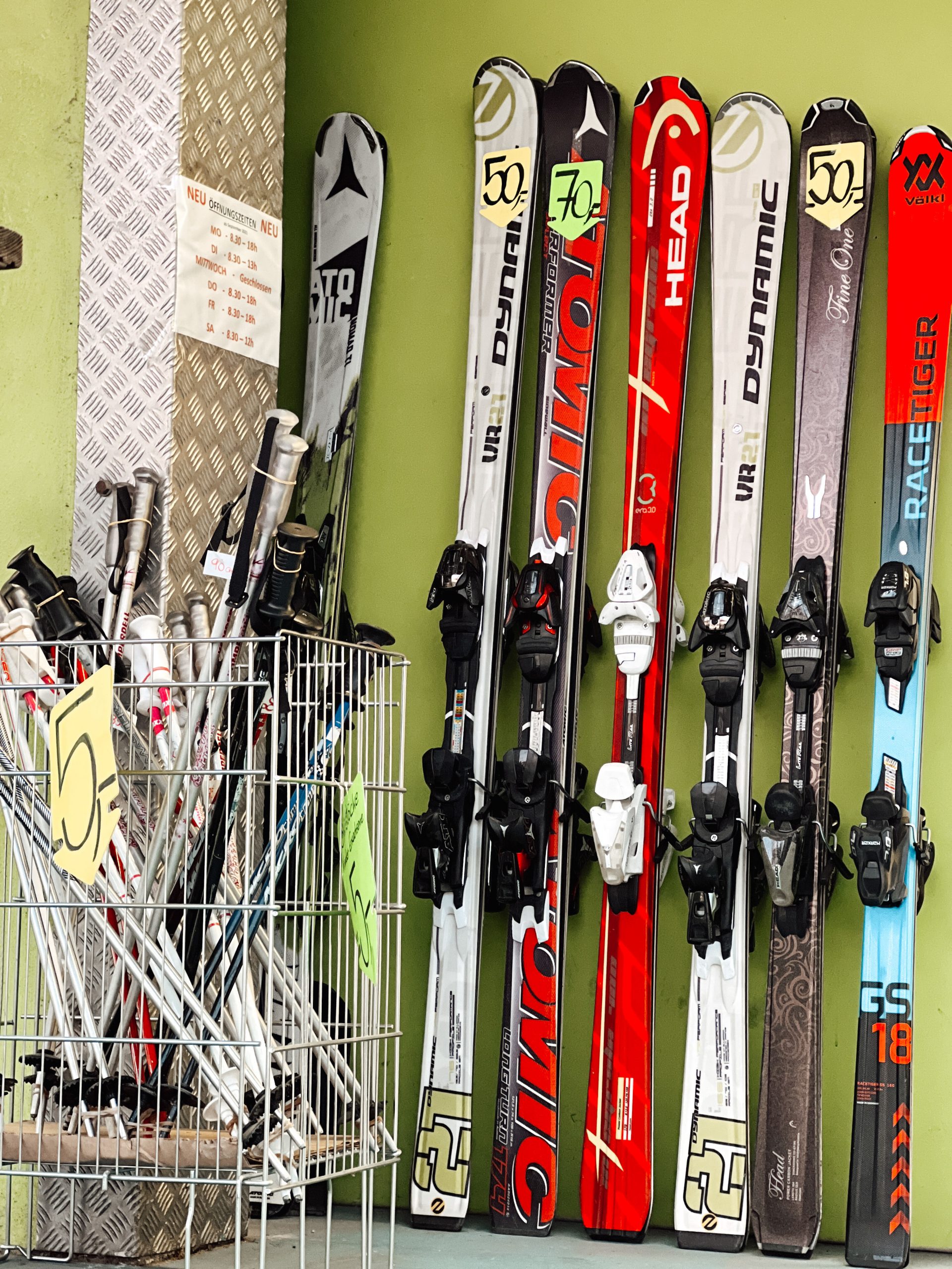 mehrere Paare Skier in einer Reihe an einer Wand, davor ein Korb mit verschiedenen Skistöcken