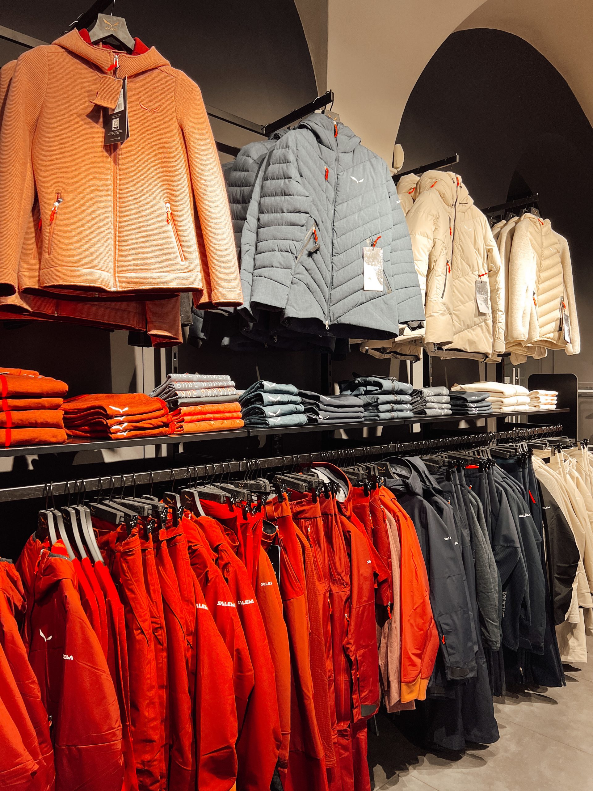 mehrere Jacken und Skibekleidung in den Farben rot, orange, ocker, grau und beige sowie dunkelblau auf Kleiderstangen (eine paralell zur Wand unten und vier normal zur Wand oben) in einem Geschäft