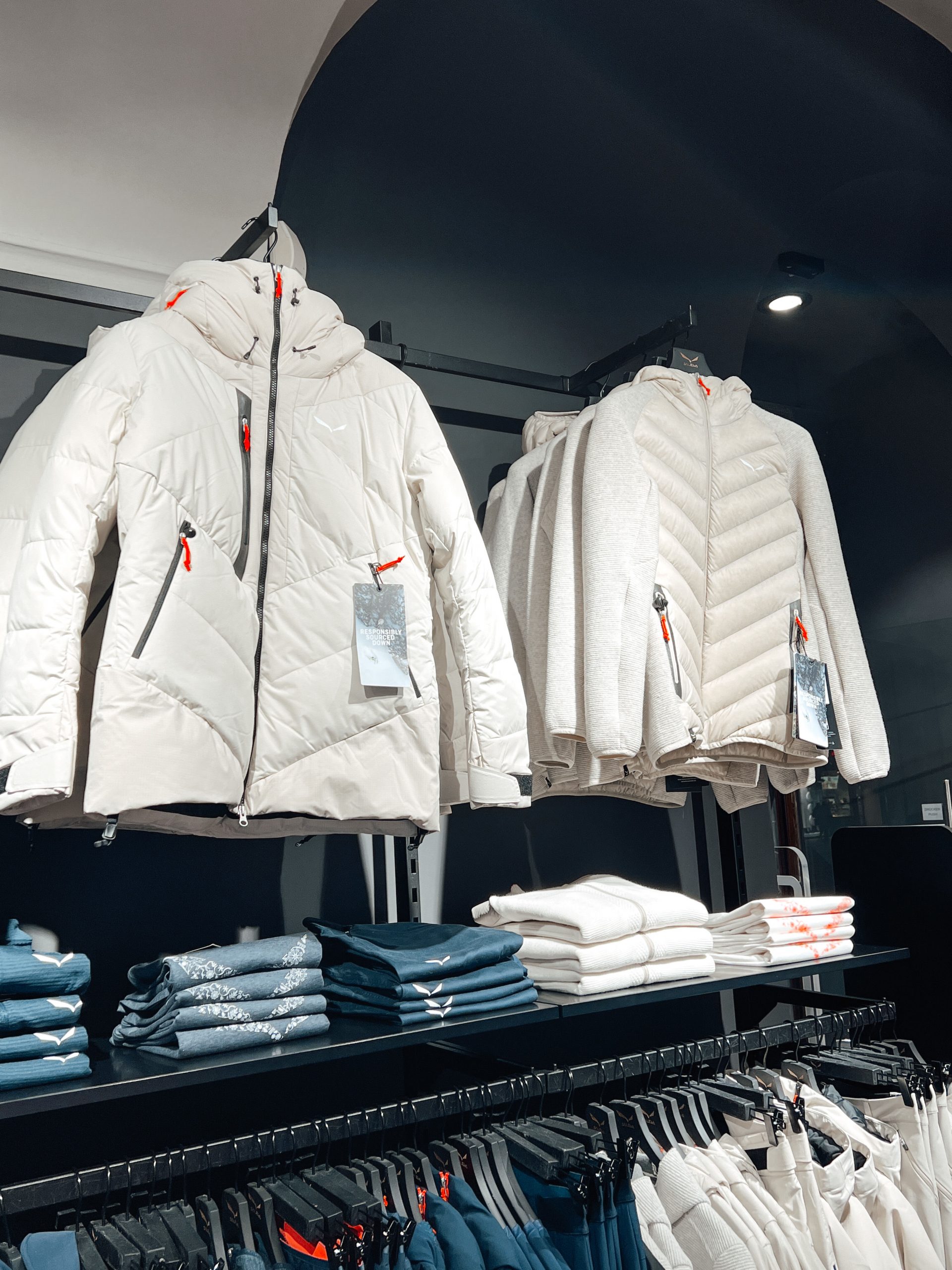mehrere weiße Jacken und weiße sowie blaue Skibekleidung auf Kleiderstangen in einem Geschäft