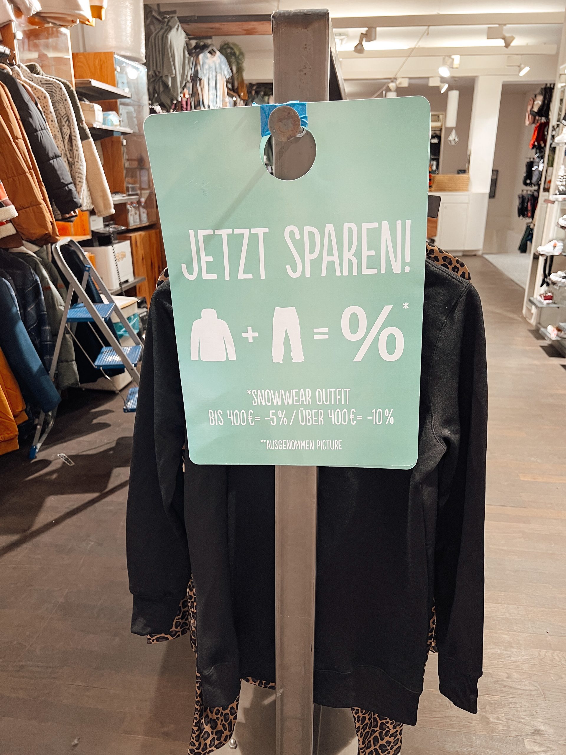Kleiderständer mit einem seitlich angebrachten Schild mit der AUfschrift "Jetzt sparen! Snowwear Outfit bis 400€ minus 5%, über 400€ minus 10%. Im Hintergrund weitere Regale in einem Geschäft